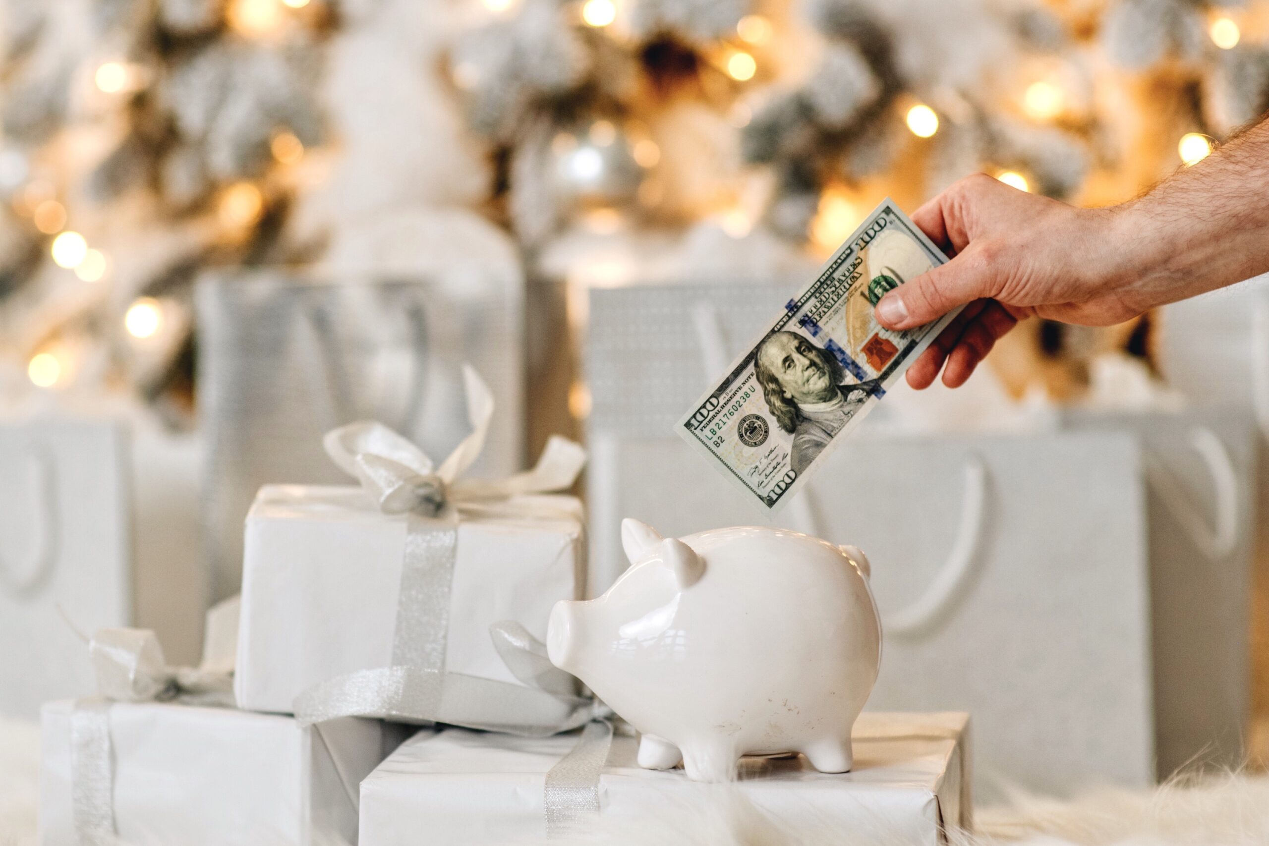 A hand places a hundred-dollar bill into a white piggy bank beside wrapped gifts and a festive background.