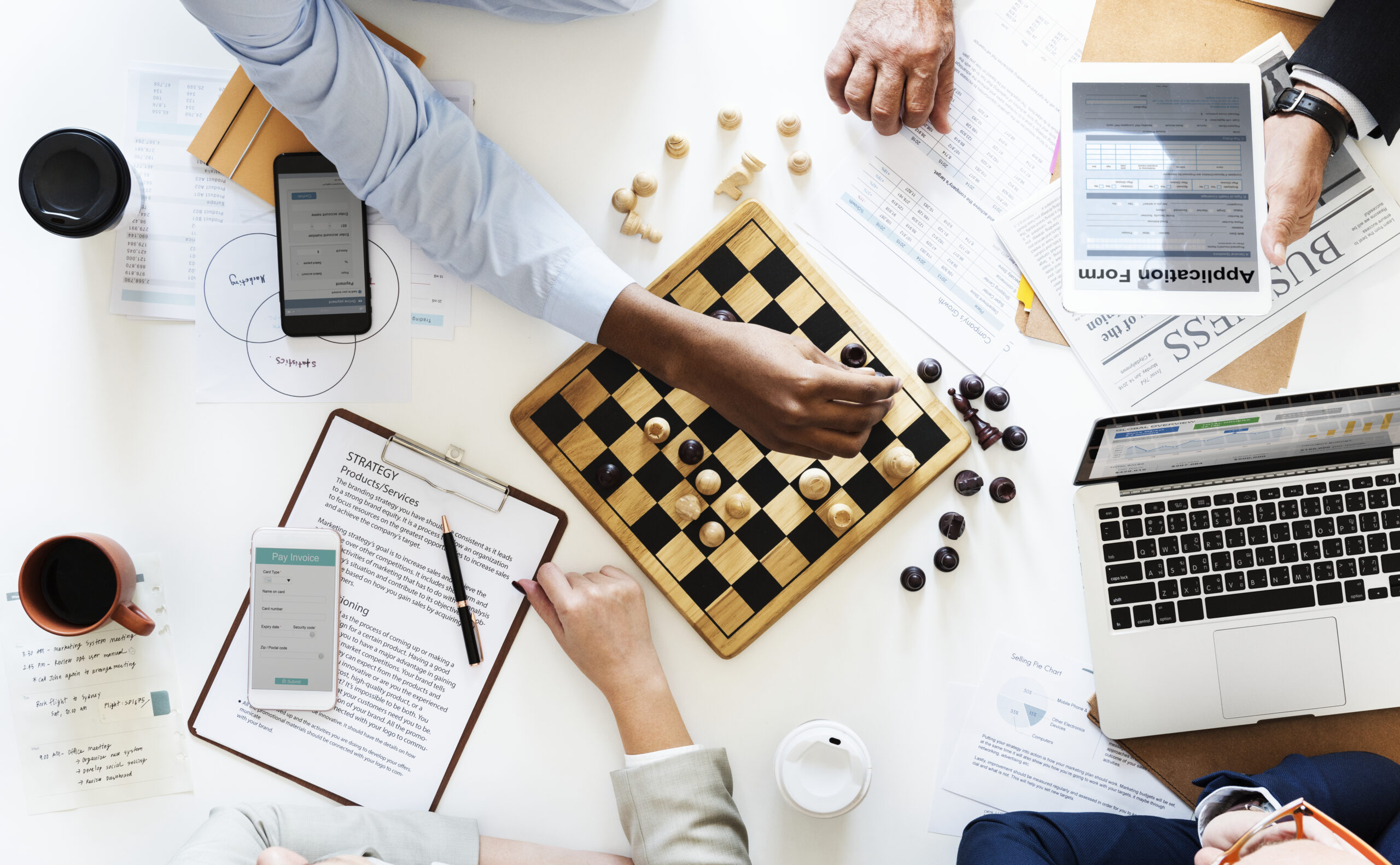 Hands engaged in a chess game on a table filled with documents, a laptop, and a smartphone.