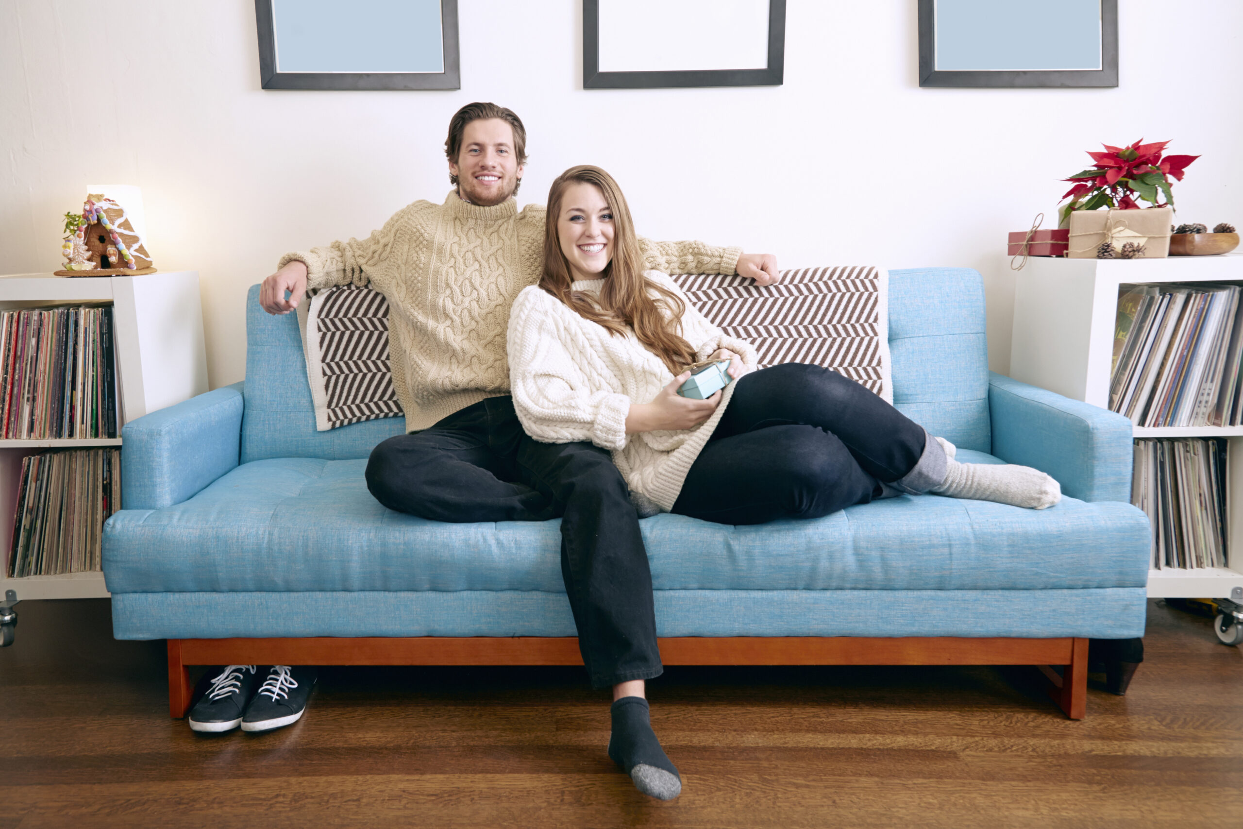 A couple in cozy sweaters relaxes on a blue couch, enjoying a moment together with a small gift.