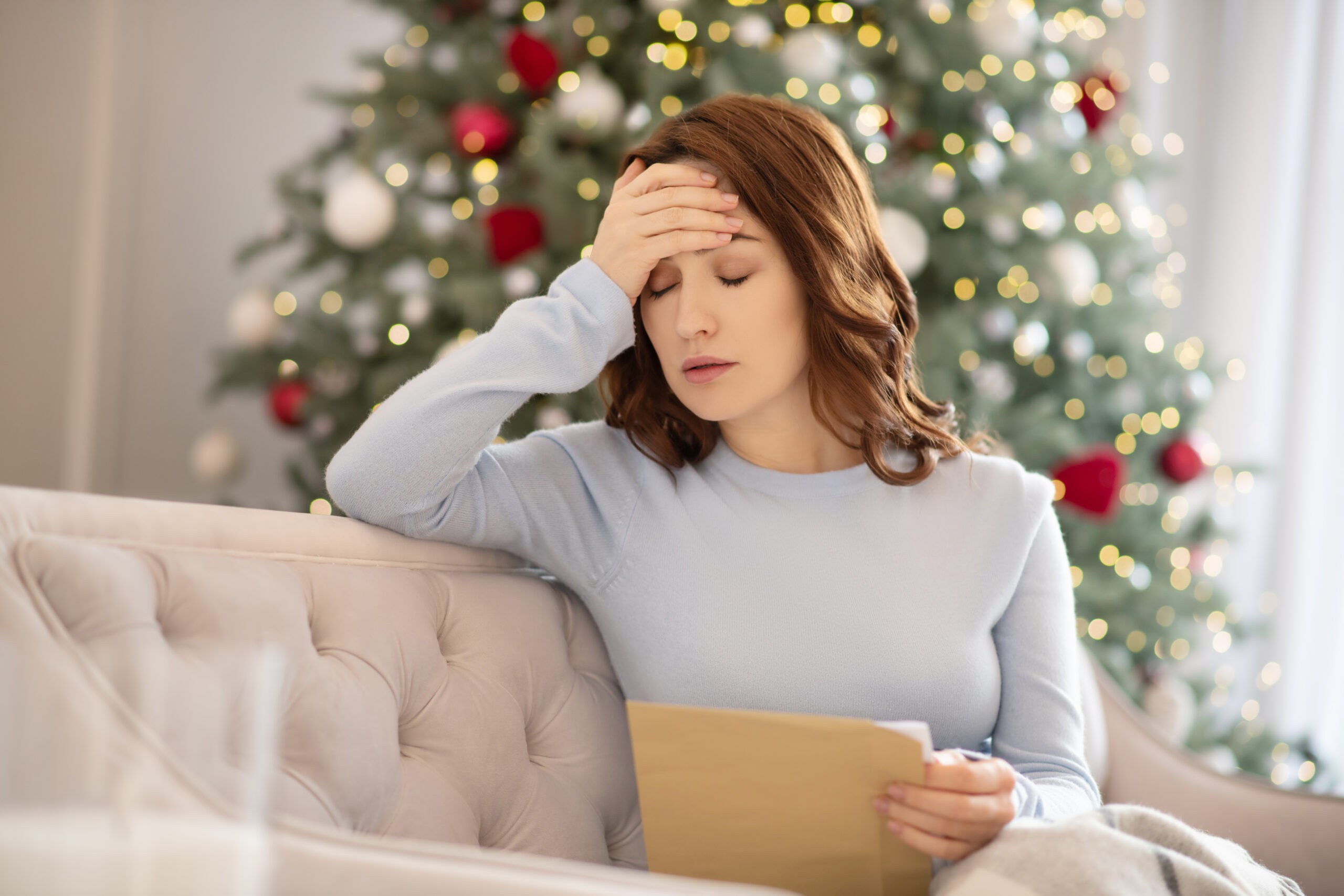 A woman sits on a couch, looking stressed while holding a letter, with a decorated Christmas tree in the background.