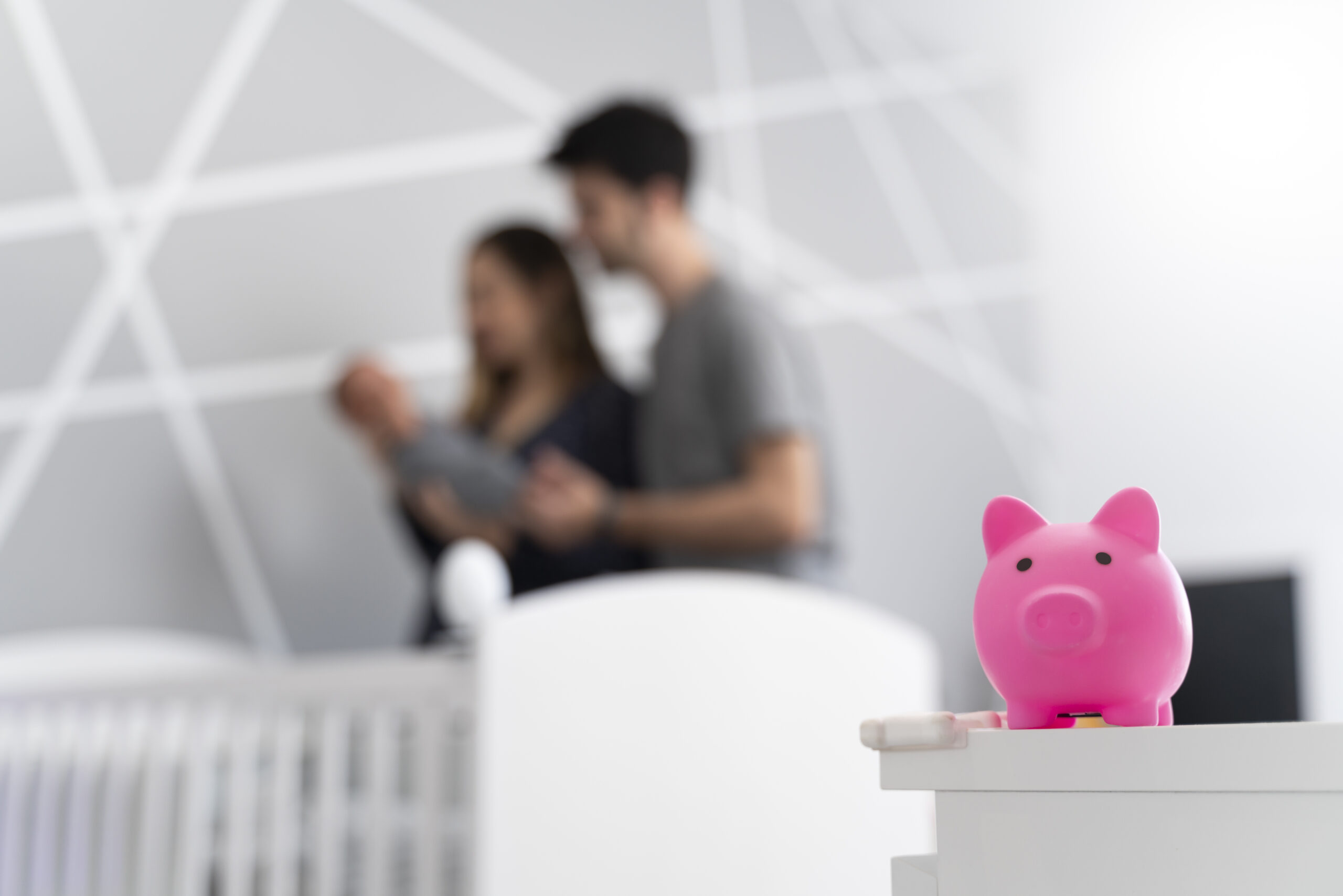A pink piggy bank sits on a dresser while a couple holds their baby in a softly lit nursery.