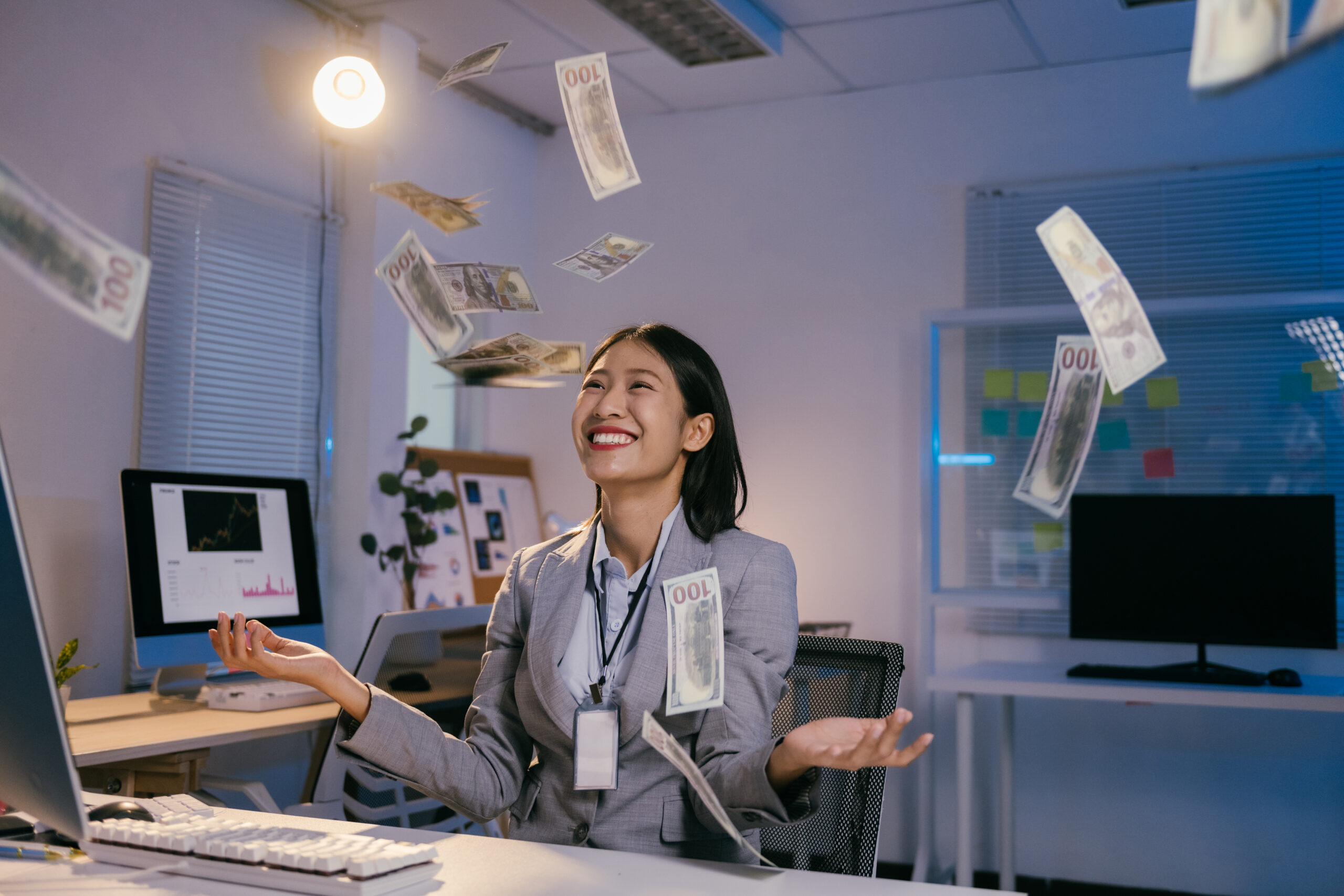 Businesswoman joyfully celebrating with flying dollar bills in a modern office setting.