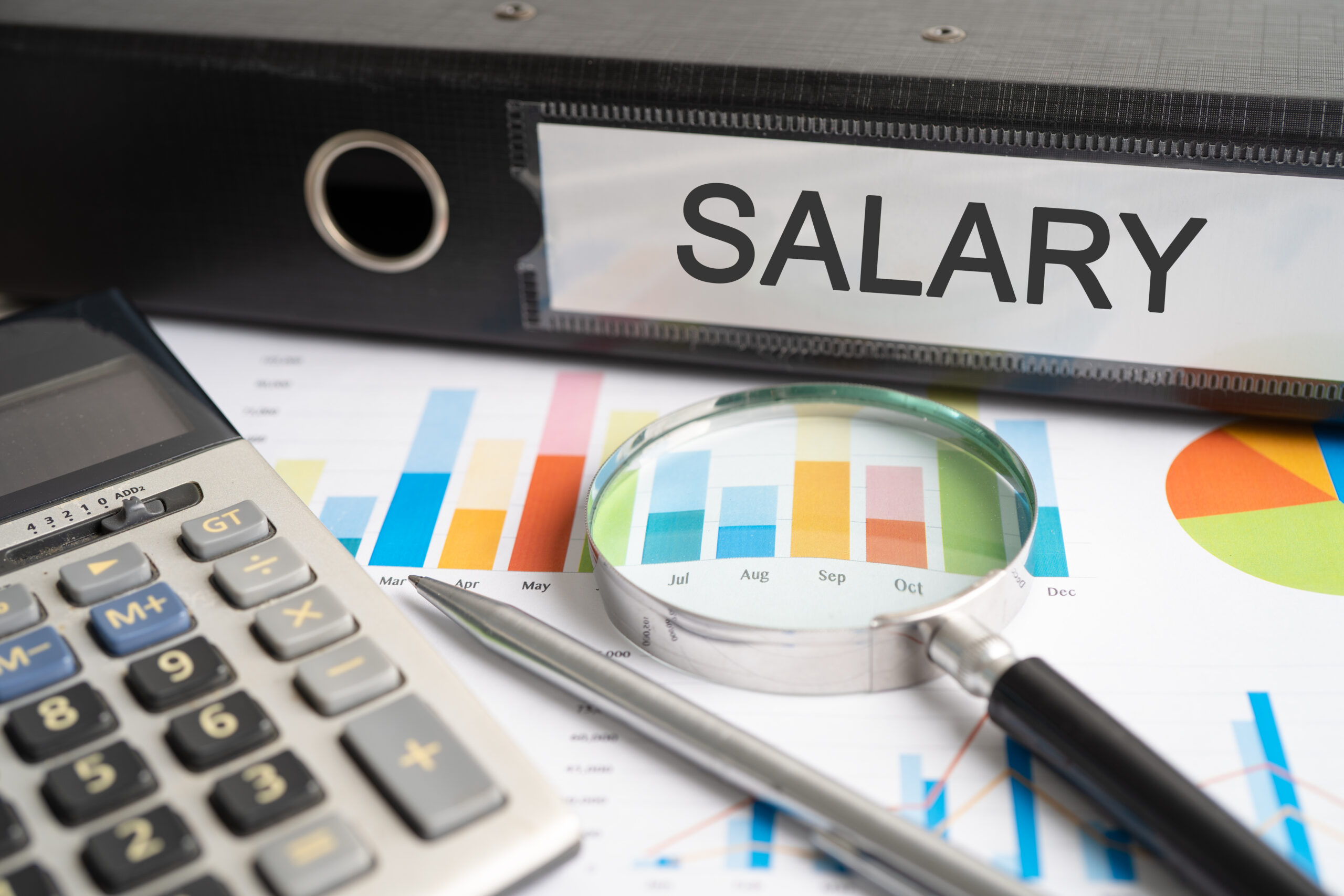 Calculator, magnifying glass, and salary binder on a financial report with colorful bar graphs and pie charts.
