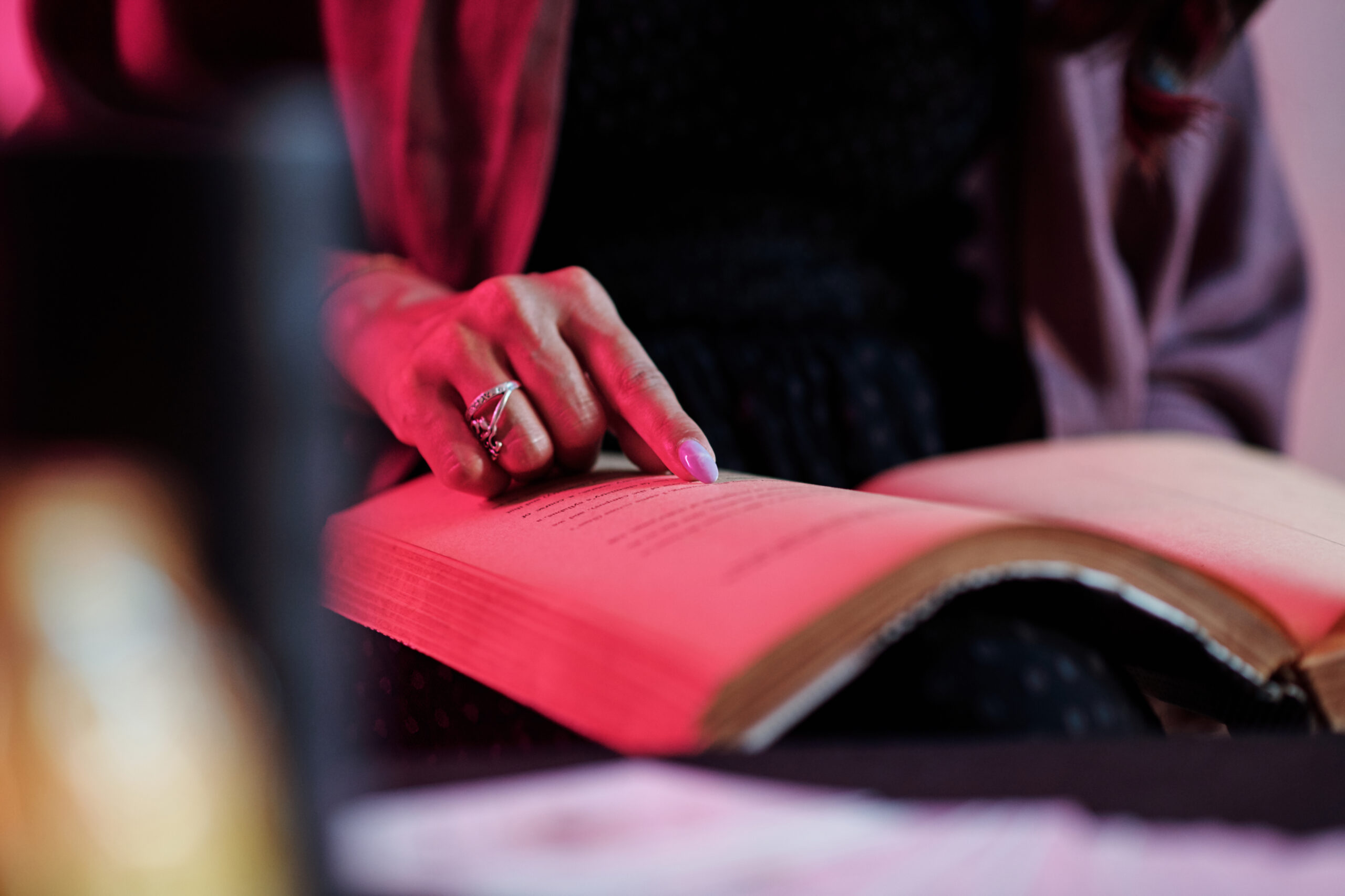 A hand with a ring points to text in an open book, illuminated by soft pink lighting.