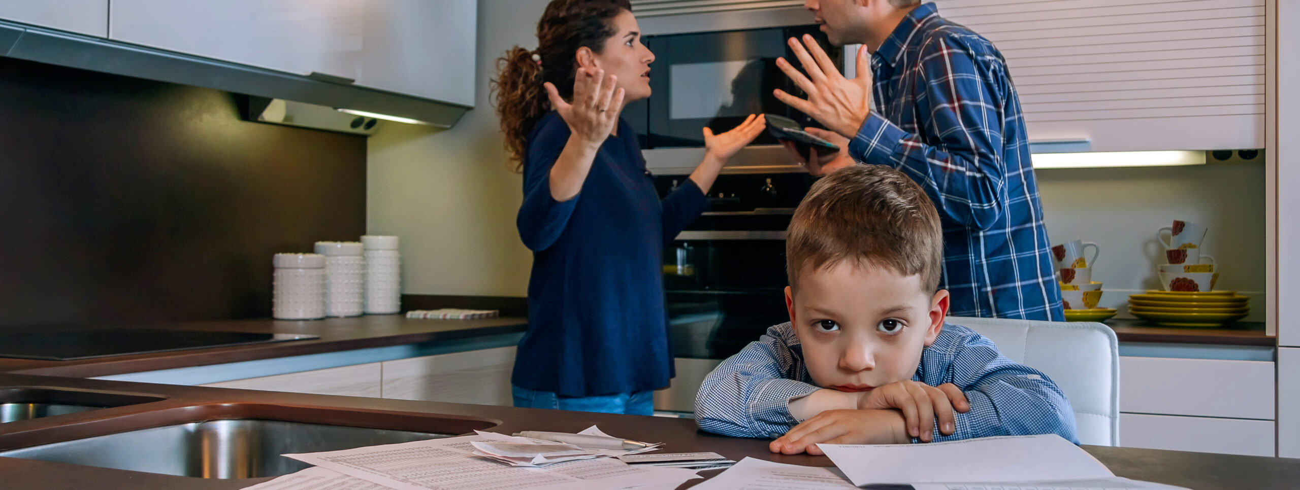 A young boy looks distressed at a kitchen table while his parents argue in the background.