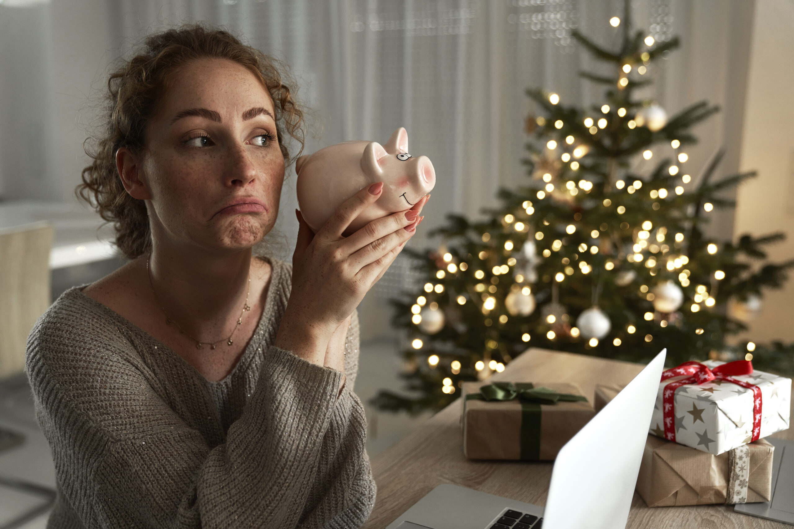 Woman with curly hair looks disappointed while holding a piggy bank, with a decorated Christmas tree in the background.