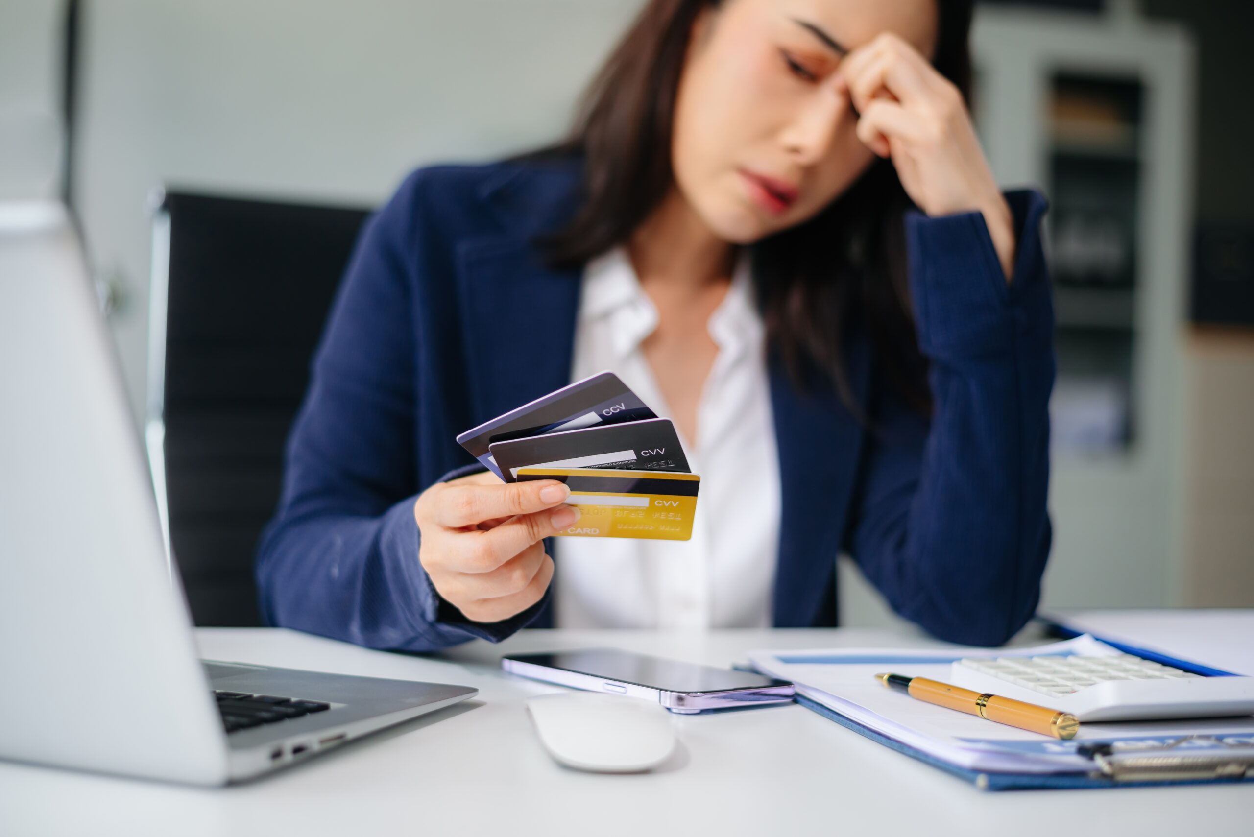 A woman looks stressed while holding multiple credit cards at a desk with a laptop and paperwork.