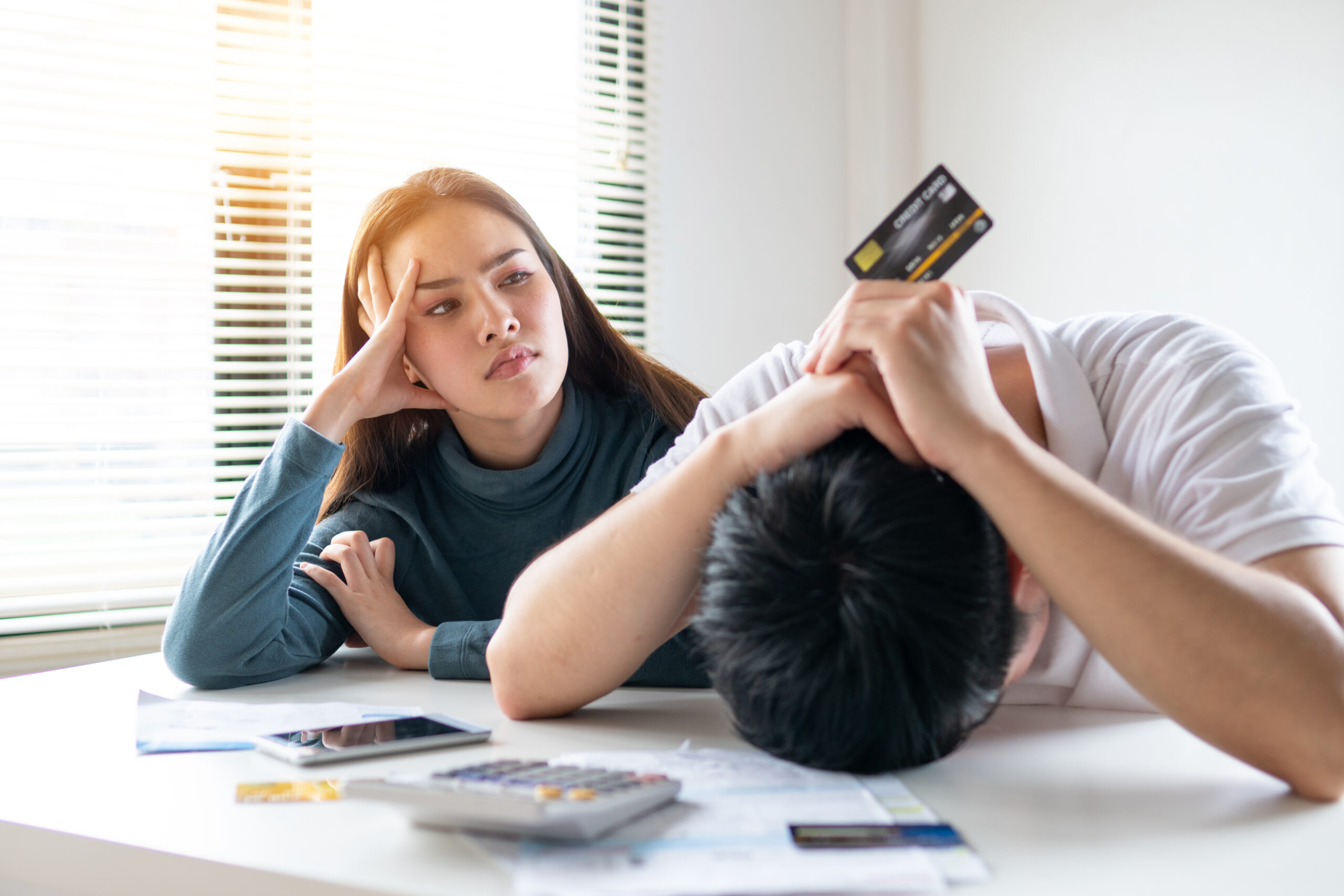 A woman looks concerned while a man with a credit card rests his head on the table, surrounded by bills and a calculator. use a loan for debt consolidation