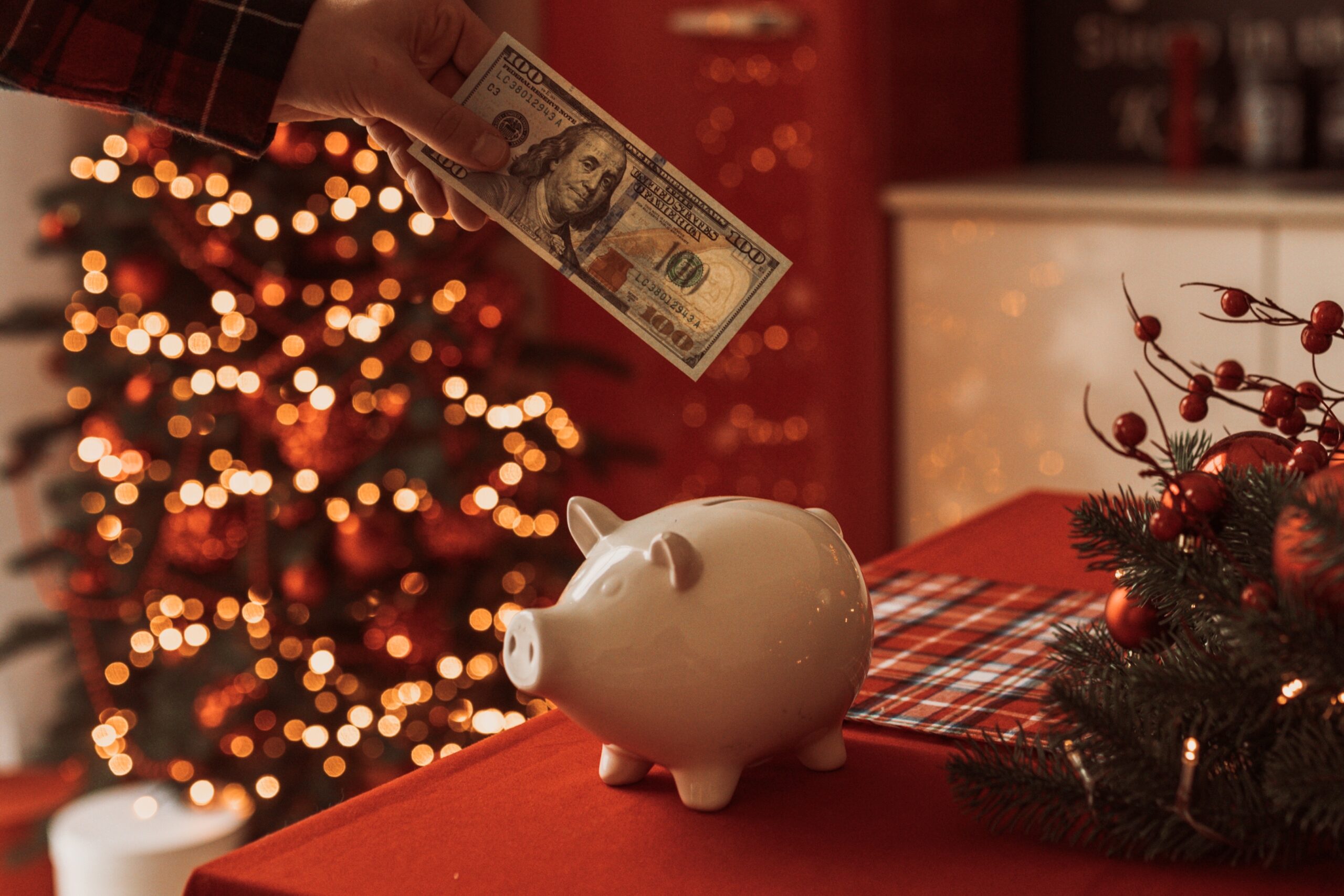 A hand places a hundred-dollar bill into a white piggy bank on a festive table with a Christmas tree in the background.