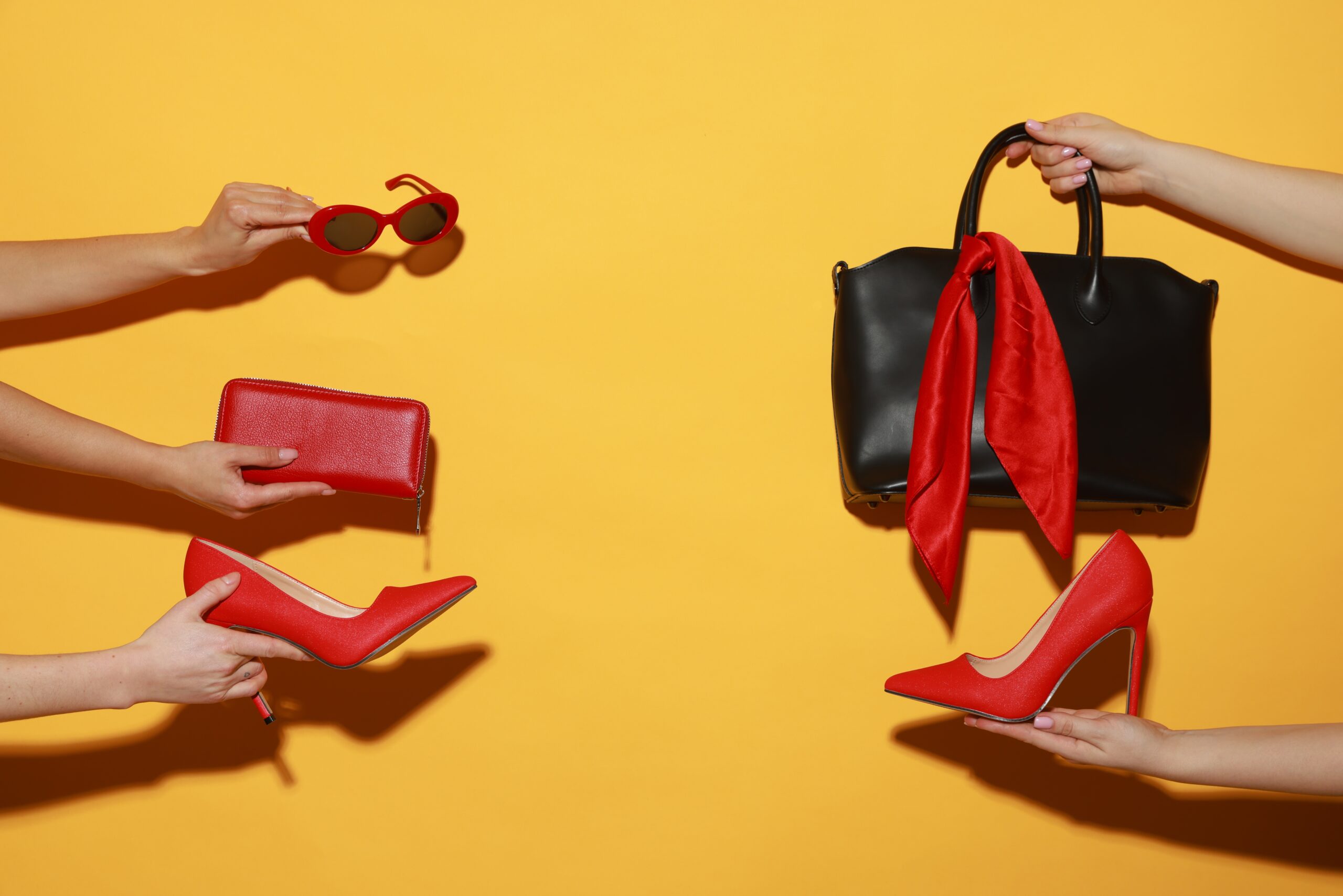 Red high heels, sunglasses, a wallet, and a black handbag with a red scarf against a bright yellow background. fake it til you make it with a luxury lifestyle